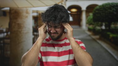 Güneşli bir günde sakallı, gözlüklü ve çizgili gömlekli genç adam endişeli ve düşünceli bir ifadeyle stresli görünüyor..
