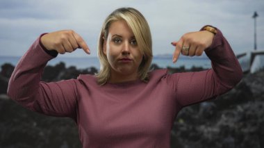 Pembe bluzlu sarışın kadın, iki eli de kayalık deniz kenarındaki denize işaret ediyor. Kararlılık..