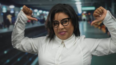 Tren istasyonunda kendinden emin bir şekilde gülümseyen, kendini gösteren başparmaklarıyla işaret eden, gözlük ve beyaz gömlek takan, kapalı alanda kendini ifade etme gücünü yakalayan bir kadın..