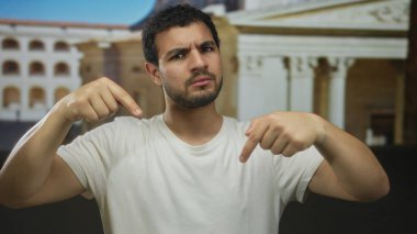Beyaz tişörtlü adam mavi stüdyoda işaret parmaklarını sıkıca aşağıya doğru yönlendiriyor..