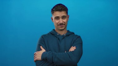 Kapüşonlu genç İspanyol adam kollarını izole edilmiş mavi duvara yaslıyor. Rahat ve rahat davranışlar sergiliyor.