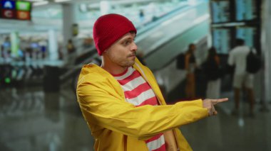 Havaalanı terminalinde sarı bir yağmurluk ve kırmızı bir bere takmış, kapalı bir alanda hareketli hareketler yapan genç İspanyol bir adam..