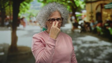 Gri saçlı yaşlı kadın şehir sokaklarında ihtiyatlı bir jest yapıyor, şehir manzarasının ortasında dikkat çekiyor..