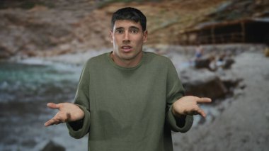 İspanyol adam sıcak güneş ışığı altında kayalık plajda ellerini açar. Şaşırmış omuz silkme hareketi gösterir. Belirsizlik..