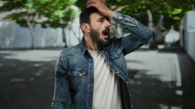 Kot ceketli İspanyol adam arka planda ağaçlarla dolu bir sokakta dikiliyor, şehir ortamında merak ve şaşkınlığını dile getiriyor..