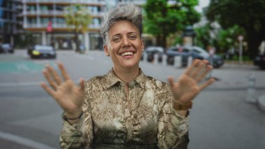 Şehir caddesinde, park halindeki arabaların yanında el çırpan kadın ve kısa saçlı yılan desenli bluz giyen bina; neşe..