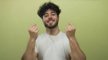 Sakallı genç İspanyol adam parmaklarıyla canlı sarı bir arkaplan karşısında kalp hareketi yapıyor. Günlük tişörtle neşe ve pozitifliği ifade ediyor..