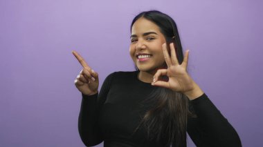 Gülümseyen bir kadın mor arka planda siyah uzun kollu bluz giyerek iyi bir hareket yapıyor..