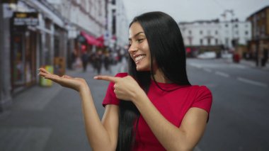 Kırmızı tişörtlü genç İspanyol kadın hareketli bir şehir caddesinde olumlu duygular ve açık havada kendine güvenen vücut dilini gösteriyor..