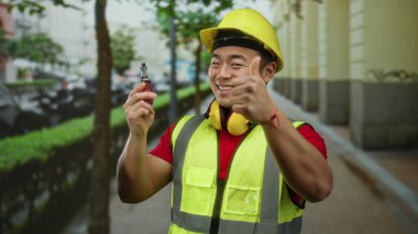Güvenlik teçhizatı içinde genç Asyalı inşaat işçisi elinde bir ses cihazı tutarken gülümsüyor, modern yaşam tarzını ve şehir ortamını ön plana çıkarıyor..