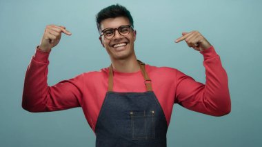 Gözlüklü ve önlüklü bir adam izole edilmiş mavi arkaplanı işaret ediyor..