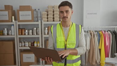 Yansıtıcı yelek giyen genç bir adam bir hayır kurumunda dizüstü bilgisayar üzerinde çalışıyor. Etrafı yiyecek ve giysi dolu raflarla çevrili. Gönüllü faaliyetlerde bulunuyor..