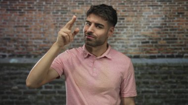 Pembe tişörtlü genç İspanyol adam stüdyoda tuğla duvara parmak işareti yapıyor; düşünceli yansıma..
