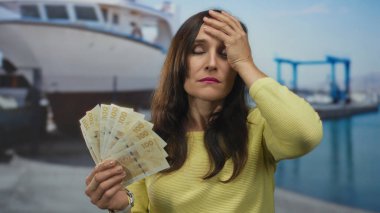 İsveçli bir kadın rıhtımda bir teknenin yanında para tutuyor, açık deniz kenarında dururken çeşitli duygular ifade ediyor..