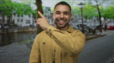 Bronz ceketli adam, bisiklet ve ağaçlarla kaplı kaldırım taşı caddesindeki kanalın kenarını işaret ediyor..