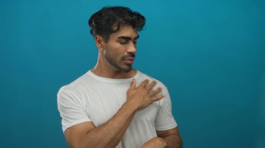 Genç İspanyol adam, mavi stüdyo ortamında beyaz gömleği göğsünün üzerinde iki eliyle ayarlar..