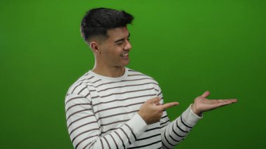 Genç adam neşeli bir şekilde göz kırpan bir ifadeyle izole edilmiş, çizgili bir süveterle yeşil bir arka plana işaret ediyor..