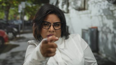 Kadın şehir sokaklarında şaşkınlık gösterirken şüpheyle işaret ediyor, gözlük ve beyaz gömlek takıyor, inançsızlık ve duygu ifadelerini yakalıyor..