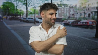 Beyaz gömlekli adam, güneşli şehir caddesinde, kanal ve binaların yanında sağ omzunu tutuyor. Huzur..