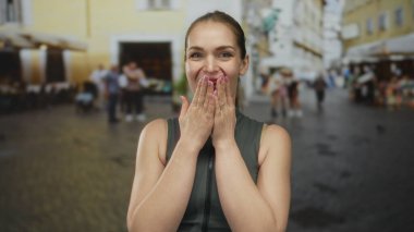 Geniş gözlü kadın, şehir merkezinde bulanık dükkanlarla dolu güneşli bir kaldırım taşı caddesinde elleri olan ağzını kapatıyor..