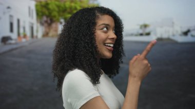 Beyaz tişörtlü kadın, kentteki beyaz binalar ve arkasında yeşil ağaçlar olan caddeleri işaret ederken, iyimser bir şekilde gülümsüyor..