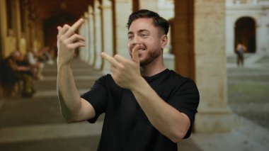 Sakallı genç bir adam eski bir üniversite koridorunda el kol hareketleri yaparak modern ifade ve klasik mimari arasındaki zıtlığı yakalıyor..