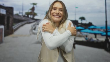 Deniz kenarındaki bir yerde gülümseyen ve kendini kucaklayan bir kadın, bulanık arka planda mavi şemsiyeler ve plaj ortamı rahatlamayı öneriyor..