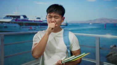 Sırt çantalı genç adam deniz kenarında öksürüyor arka planda teknesi olan kitapları açık gökyüzünün altında açık hava öğrenciliğini simgeliyor..
