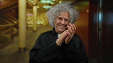 Bir otel lobisinde el çırpan, sevinç ifade eden, yazman kıyafeti giyen, canlı bir gülümsemesi olan, gri saçlı, kapalı bir ortamda olgun, sıcaklığını ve mutluluğunu ifade eden bir kadın..