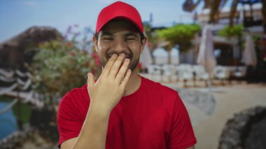 Kırmızı gömlekli ve şapkalı adam, sokak restoranının terasında güneş ışığı altında ağzını eliyle kapatıyor..