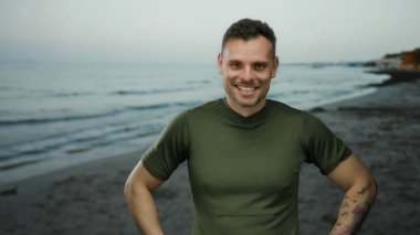 Gün batımında sakin bir plajda dururken güvenle gülümseyen dövmeleri olan genç bir adam. Okyanus dalgaları, berrak bir gökyüzünün altında yavaşça arka planda geziniyor..