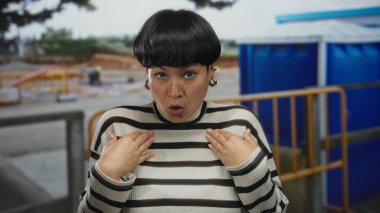 Çizgili kazaklı genç Çinli kadın, ağaçlardan ve mavi çitlerden oluşan bir gölgeliğin altında açık hava şantiyesinde belirgin bir şekilde el kol hareketi yapıyor..