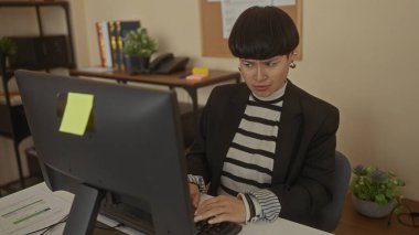 Ofis ortamında bilgisayar başında çalışan bir kadın, masasının etrafında bitki ve ofis malzemeleriyle modern bir iş ortamında odaklanmış ve niyetlenmiştir..