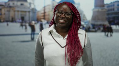 Vatikan açıkhava meydanında kırmızı örgülü, gözlüklü ve beyaz gömlekli gülümseyen kadın, tarihi kent ortamında neşeli ve canlı bir anı yakalıyor..