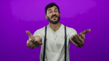 Sakallı İspanyol bir adam canlı mor bir arka plana karşı davetkar bir şekilde hareket ediyor beyaz gömlek ve pantolon askısı giyiyor arkadaşlığını ve sıcaklığını açık kollarla ifade ediyor.