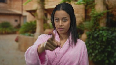 Pembe bornozlu kadın, sakin bir açık hava ortamında düşünceli bir şekilde el kol hareketi yapıyor, yemyeşil bitkiler ve kırsal mimariyle çevrili rahat ve rahat bir atmosferi vurguluyor..