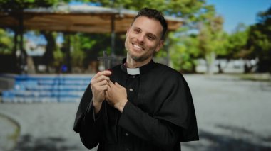 Siyah cüppeli Katolik bir rahip elinde bir haç, açık hava parkı ortamında, güneş ışığı altında huzur ve yansıma simgeliyor..