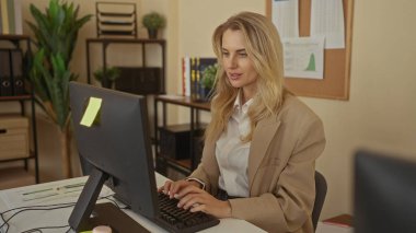 Modern işyerindeki bilgisayar masasında çalışan, sarı saçlı, gülümseyen, daktilo kullanan ve arka planda bej dekoru ve bitkileri olan bir kadın..
