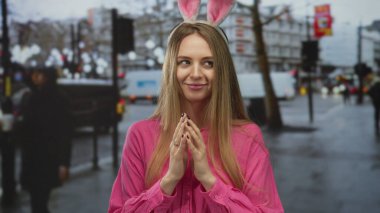 Şehir arka planında pembe giyinmiş bir sokakta tavşan kulağı takan bir kadın hareketli bir ortamda neşeli paskalya titreşimleri yayıyor..