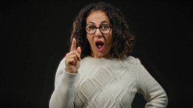 Kıvırcık saçlı gözlüklü İspanyol kadın şaşkınlığını dile getiriyor ve siyah arka planda bir fikir belirterek parmağını kaldırdı..