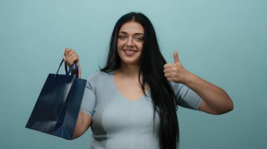 Uzun saçlı, mavi yalıtılmış arka planda poz veren kıvrımlı genç bir kadın, mavi bir alışveriş çantası tutarken baş parmaklarını gösteriyor, karışık duygular ifade ediyor..