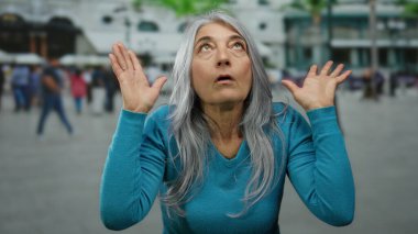 Gri saçlı bir kadın, kalabalık bir şehir caddesinde, açık havada, insanlarla çevrili, şehir ortamında duygu ve titreşim sergiliyor..