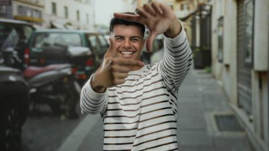 Çizgili tişörtlü genç adam gülümsüyor ve elleri ile şehir dışında canlı bir sokak arkaplanında çerçeveleniyor.