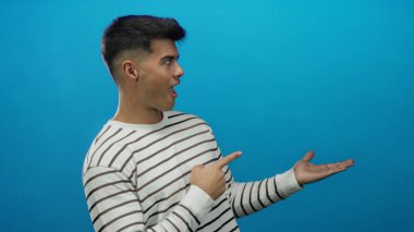 Çizgili gömlekli genç adam gülümsüyor ve mavi arka plana işaret ediyor.