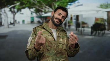 Askeri üniformalı İspanyol bir adam arka planda ağaçlarla güneşli bir sokakta para hareketi yapıyor..