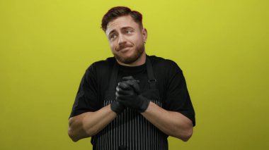 Önlük ve eldiven takan sakallı genç adam parlak sarı arka planda duruyor ve neşeli bir ifadeyle omuz silkiyor..