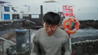 Gri kazaklı genç adam sahilde duygularını ifade ediyor. Arka planda can yelekleri ve binalar var. Sahilde bir açık hava sahnesi sergiliyor..