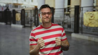 Çizgili tişörtlü genç adam başparmağını kaldırıyor ve kent geçmişiyle gülümsüyor. Şehir sokaklarında pozitif ve kendine güvenini gösteriyor..