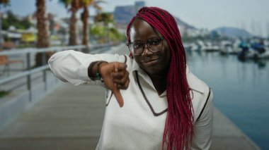 Örgü örgülü bir kadın açık havada açık bir gökyüzünün altında botları ve palmiyeleri olan bir limanda baş parmağını sallıyor..