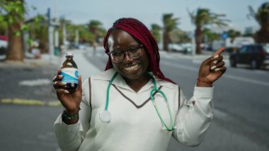 Güneşli bir caddede elinde ilaç tutarken steteskoplu gülümseyen ve puanlı Afrikalı Amerikalı kadın doktor..